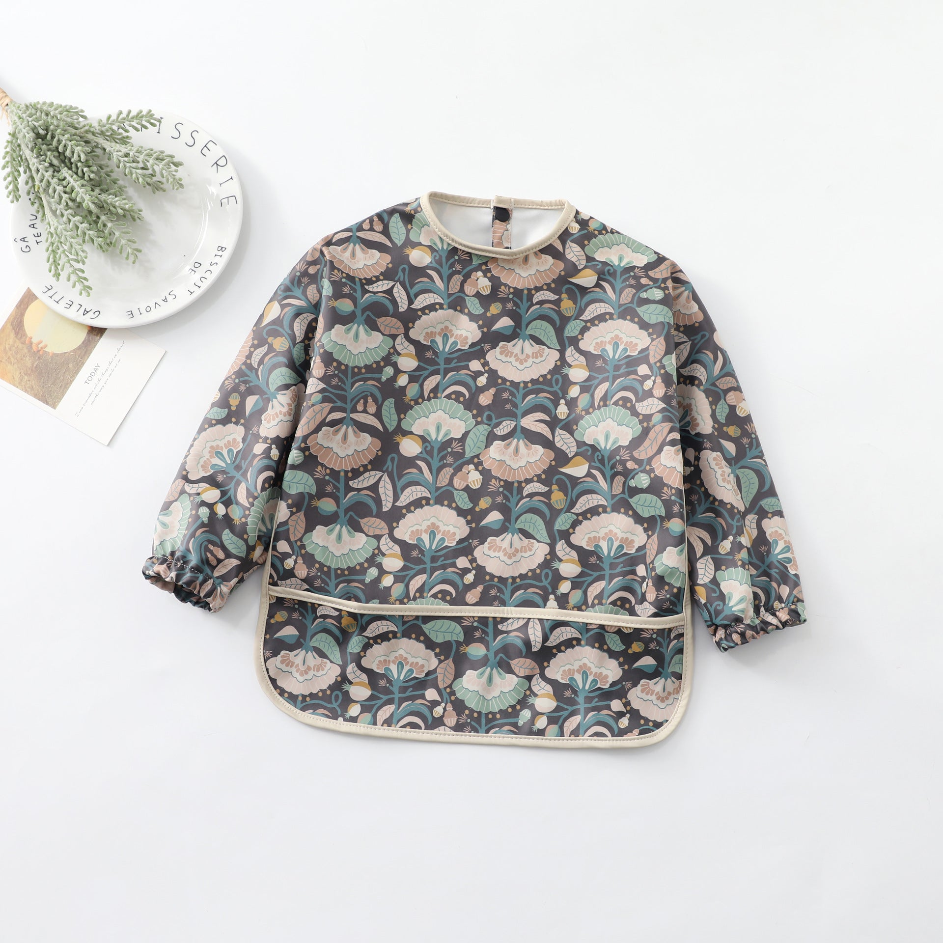 a children's bib with black background. The contents are peony flowers of cream and green colors with green stalk and leaves. The bib is shown next to a plate with a plant on it.